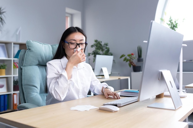 Une employée de bureau asiatique malade a le rhume et la grippe tousse et éternue dans les tissus fonctionne à l'ordinateur