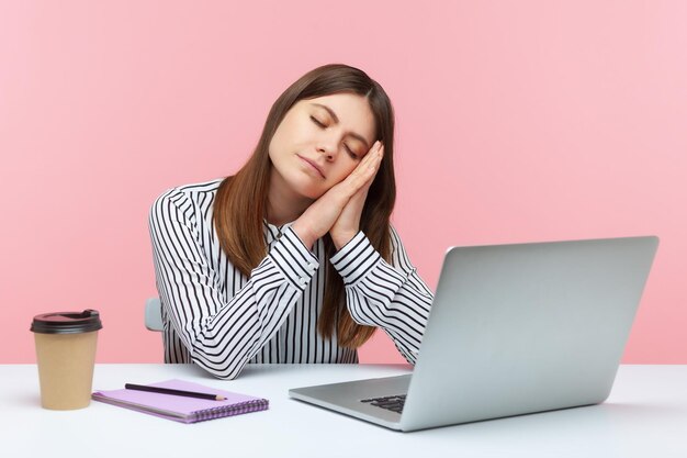 Employée de bureau endormie ennuyée en chemise rayée faisant la sieste penchée sur les mains assise sur le lieu de travail avec un ordinateur portable, surmenage, réveil précoce. Studio intérieur tourné isolé sur fond rose