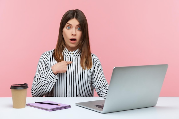 Employée de bureau femme choquée se pointant du doigt et regardant la caméra avec une expression surprise étonnée de la tâche de travail assis sur le lieu de travail avec un ordinateur portable Prise de vue en studio intérieur isolée sur fond rose