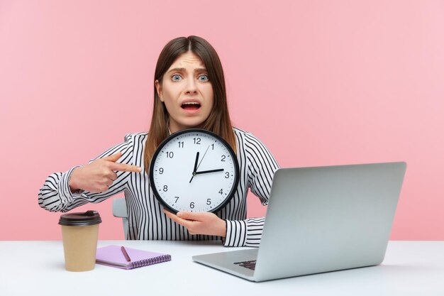 Employée de bureau nerveuse et anxieuse pointant le doigt vers une grande horloge murale dans ses mains assise sur le lieu de travail, travaillant tard, heures supplémentaires. Studio intérieur tourné isolé sur fond rose