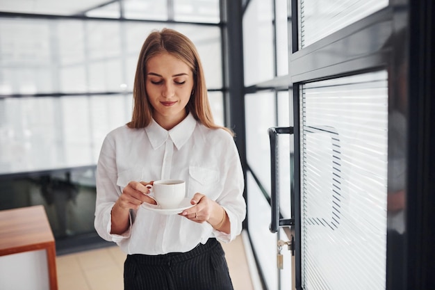 Employée de bureau en vêtements formels debout à l'intérieur pendant la journée avec une tasse de boisson.