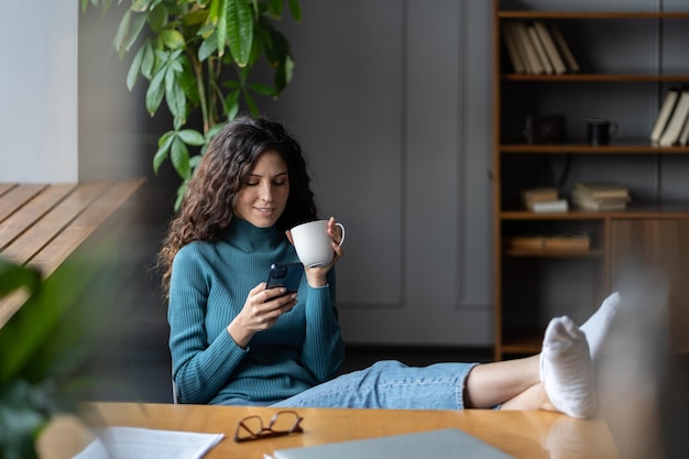 Employée détendue buvant du café et utilisant un téléphone portable sur le lieu de travail avec les jambes sur la table de bureau