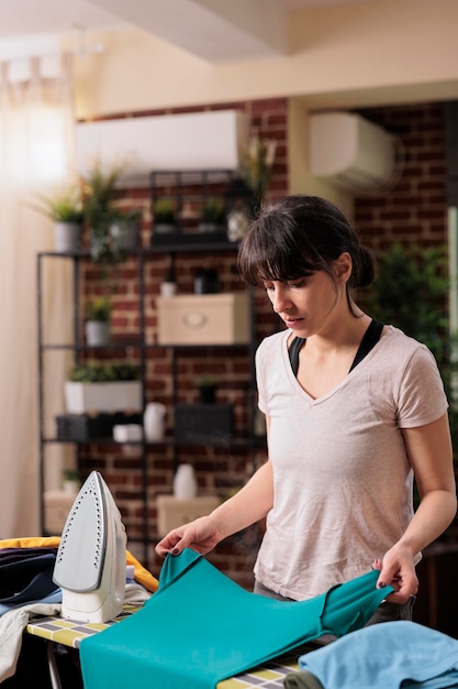 Photo employée de maison femme repassant des vêtements sur une planche à repasser dans un salon d'appartement moderne. femme concentrée faisant ses devoirs sur le lieu de travail, chargée de tout garder en ordre.