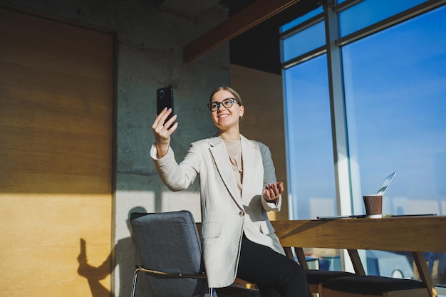 Employée positive mignonne avec de longs cheveux blonds dans des vêtements décontractés regardant le téléphone tout en travaillant sur un nouveau projet d'entreprise à table avec un ordinateur portable et des gadgets au bureau