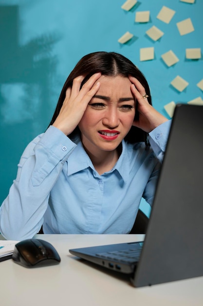 Une employée souffrant de maux de tête se frottant les tempes et ayant l'air frustrée devant l'écran d'un ordinateur portable. Femme professionnelle asiatique souffrant de migraine se sentant stressée par la charge de travail au travail
