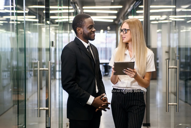 Employés de bureau africains hommes et femmes de race blanche avec tablet pc debout au couloir de bureau vide et discuter de nouveau projet.