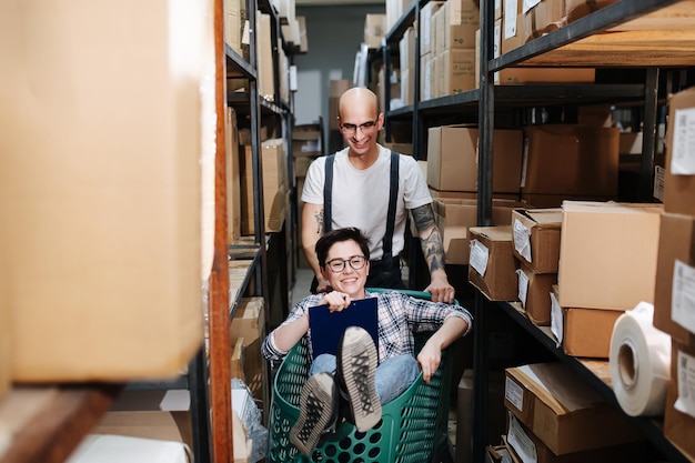 Des employés d'entrepôt souriants jouant avec un chariot qu'elle monte dedans qu'il pousse