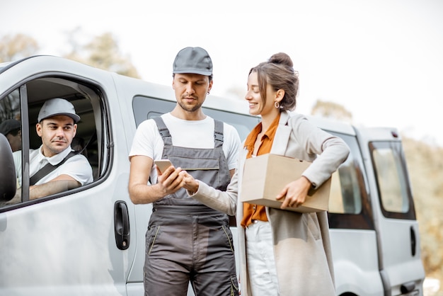 Employés de l'entreprise de livraison en uniforme livrant des marchandises à un client par véhicule fourgon, femme signant sur un smartphone, recevant un colis à l'extérieur