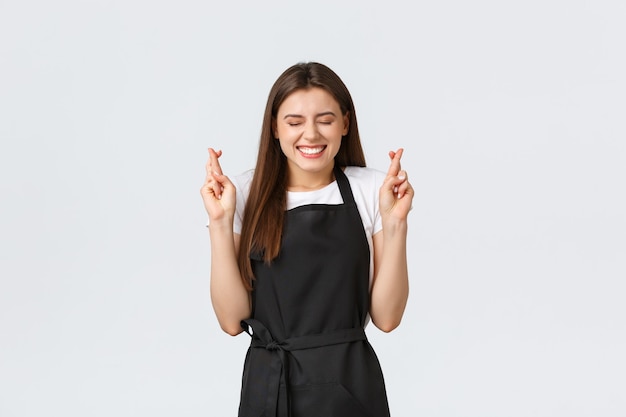 Employés d'épicerie, concept de petites entreprises et de cafés. Excitée et joyeuse belle femme barista en tablier noir souriant optimiste, croiser les doigts bonne chance, faire un vœu