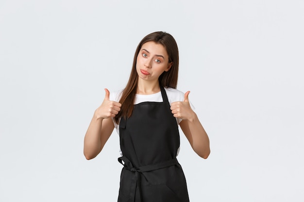 Employés d'épicerie, concept de petites entreprises et de cafés. Une femme barista enthousiaste et gaie montre son pouce levé en signe d'approbation. Vendeuse en tablier noir impressionnée par une nouvelle promo cool