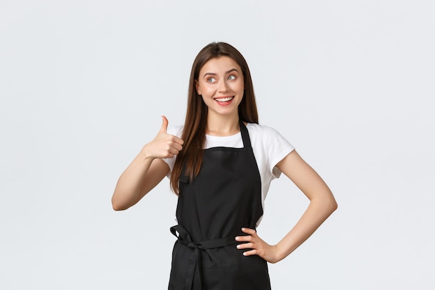 Employés d'épicerie, concept de petites entreprises et de cafés. Une femme barista satisfaite en tablier noir montre son pouce levé en signe d'approbation. La vendeuse recommande le produit. Le travailleur du café donne des conseils.