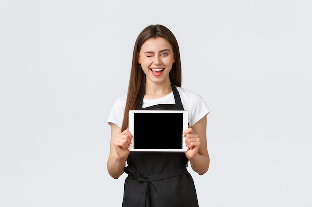 Employés d'épicerie, concept de petites entreprises et de cafés. Une femme barista souriante et excitée, une employée de café en tablier montrant l'affichage d'une tablette numérique, annonçant un nouveau menu ou une commande en ligne.