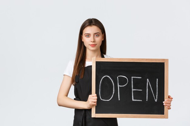 Employés d'épicerie, concept de petites entreprises et de cafés. Jolie jeune employée de magasin en tablier noir tenant une pancarte ouverte, informer les invités que le café fonctionne, fond blanc