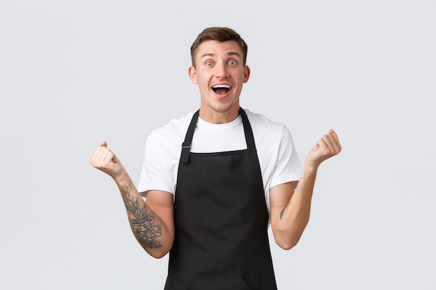 Employés, épiceries et concept de café. Heureux et soulagé souriant beau barista, serveur en tablier noir, pompe à poing en réjouissant, victoire triomphante ou bonne nouvelle positive