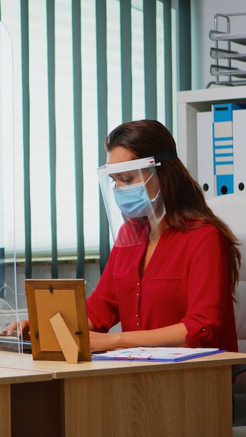 Photo employés portant un masque et une visière assis dans une nouvelle salle de bureau normale respectant la distanciation sociale. collègues travaillant dans un lieu de travail moderne respectant les règles de protection contre le virus covid à l'aide de plexiglas.