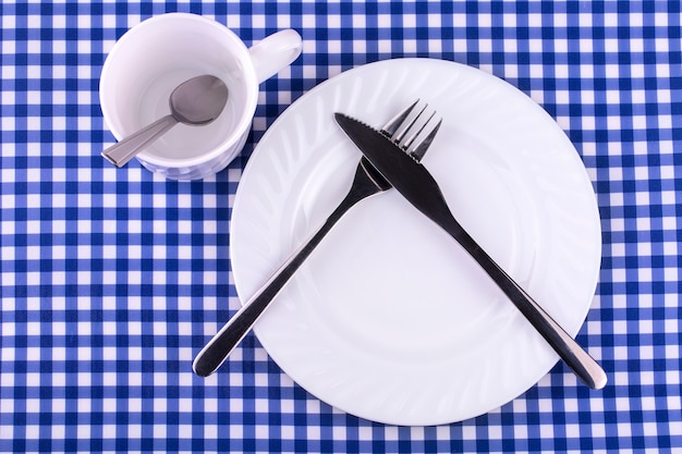 Empy Mug avec cuillère, assiette avec couteau et fourchette sur la nappe dans une cage. Vue d'en haut avec espace de copie