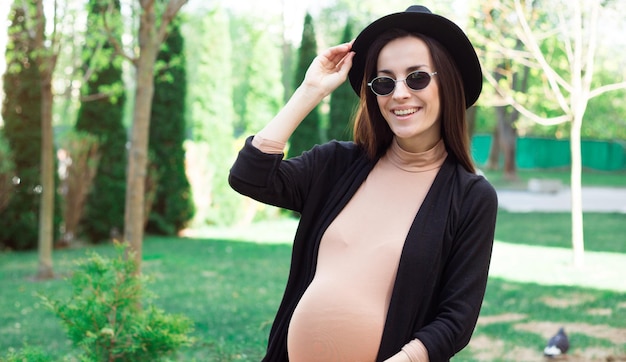 Enceinte élégante en chapeau et lunettes de soleil assise sur un banc dans le parc de la ville et se détendre