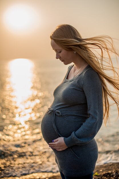 Enceinte jeune femme debout sur la plage dans une robe grise en tricot. Concept de grossesse