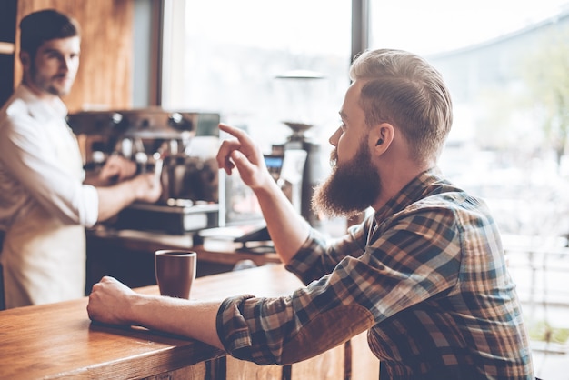 Encore un café s'il vous plait ! Jeune bel homme barbu parlant au barista alors qu'il était assis au comptoir du bar au café