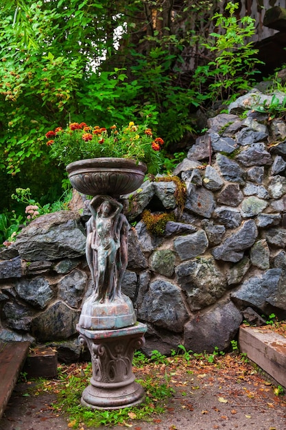 Un endroit pittoresque avec un parterre de fleurs sous la forme d'une statue de femme nue tenant un vase contre un mur de pierre parmi le feuillage vert et les arbres le jour d'été