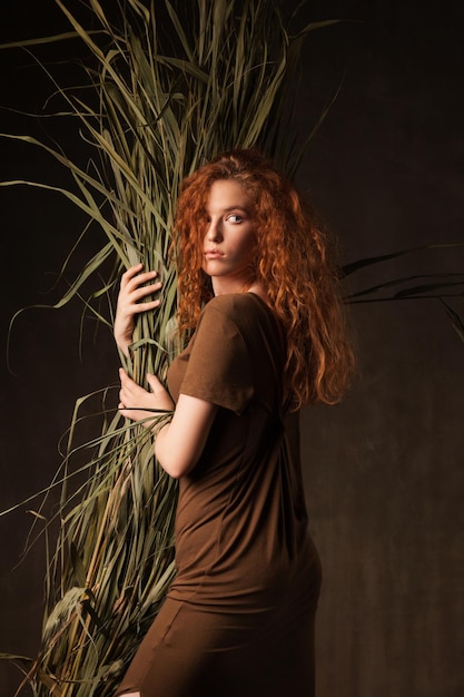 Energizer belle jeune femme aux cheveux roux bouclés avec de l'herbe de canne de plante tropicale verte posée dans des murs sombres studio Copier l'arrière-plan de l'espace