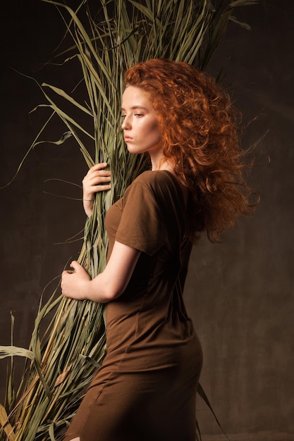 Energizer belle jeune femme aux cheveux roux bouclés avec de l'herbe de canne de plante tropicale verte posée dans un studio de murs sombres