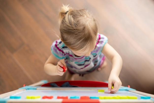 Un enfant de 2 ans se tient sur un parquet marron et dispose des numéros magnétiques sur le tableau Vue d'en haut