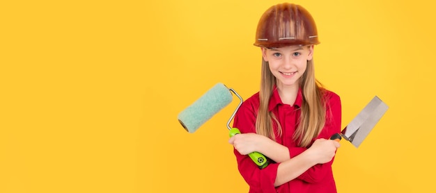 Enfant adolescent positif dans un casque de constructeur avec rouleau à peinture et spatule sur mur jaune Constructeur d'enfant dans la conception d'affiche horizontale de casque Espace de copie d'en-tête de bannière