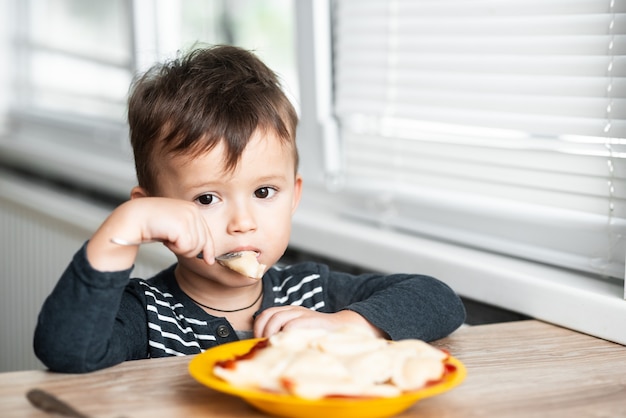 Enfant affamé mangeant des boulettes dans la cuisine, se reposant à la table dans une veste grise