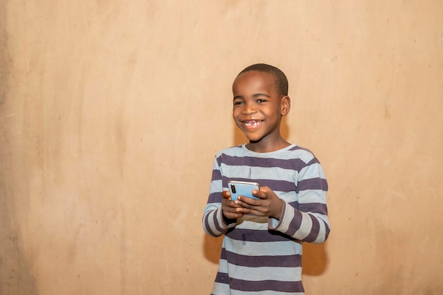 Enfant africain mignon utilisant un téléphone intelligent et souriant, debout sur un mur jaune pastel, fond