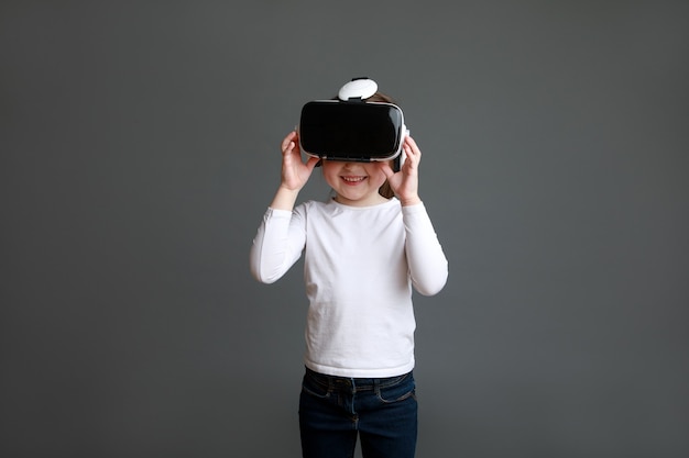 Enfant d'âge préscolaire en chemise à manches longues blanche portant des lunettes de réalité virtuelle sur une surface grise.