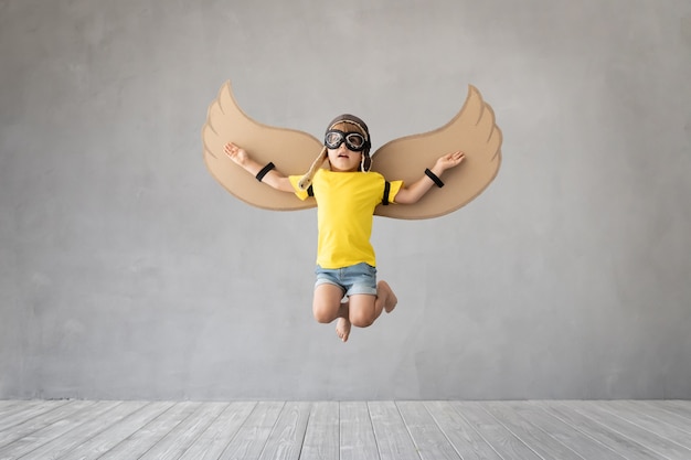 Enfant avec des ailes en papier sautant contre le mur de béton gris.