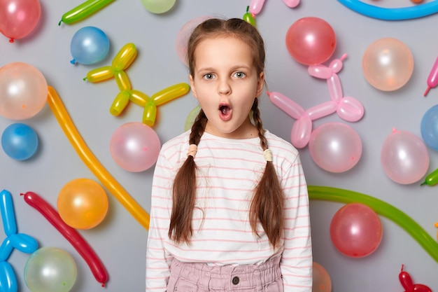 Enfant d'anniversaire étonné Petite fille surprise avec des nattes debout contre un mur gris décoré de ballons colorés regardant la caméra avec de grands yeux voit un cadeau choqué