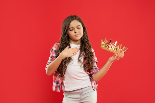 Un enfant arrogant aux cheveux bouclés tient la couronne de la reine sur fond rouge, concours