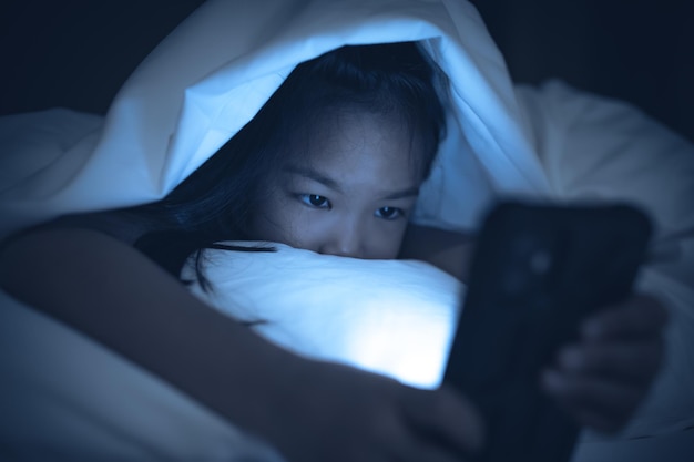 Enfant asiatique jouant au jeu sur smartphone dans le lit la nuitLa fille Addict des médias sociaux
