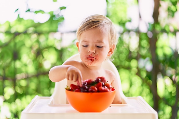 Un enfant au visage sale est assis à table et cueille des fruits dans une assiette