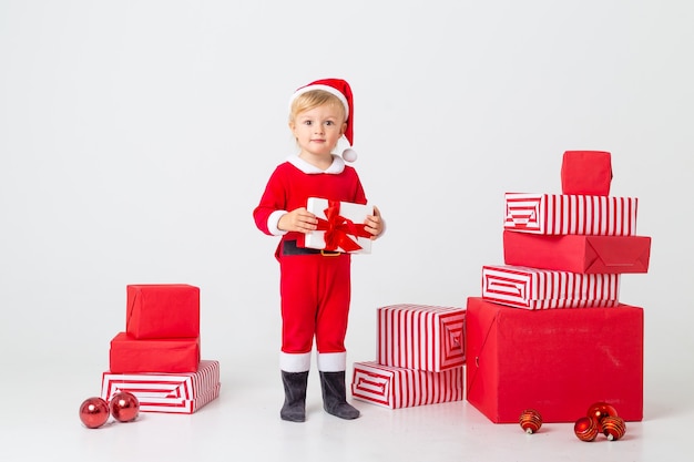 Un enfant en bas âge dans un costume de Père Noël se dresse sur un fond blanc à côté de coffrets cadeaux pour Noël. Concept de Noël, espace de texte