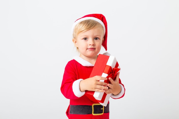 Un enfant en bas âge dans un costume de Père Noël se dresse sur un fond blanc à côté de coffrets cadeaux pour Noël. Concept de Noël, espace de texte