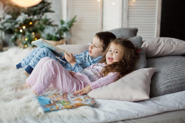 Enfant en bas âge garçon et fille en pyjama allongé sur le lit et lire des livres près de l'arbre de Noël dans l'intérieur de Noël clair