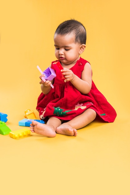 Enfant en bas âge indien ou asiatique ou nourrisson ou bébé jouant avec des jouets ou des blocs en position couchée ou assise isolé sur fond clair ou coloré