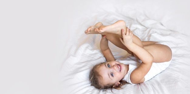 Enfant en bas âge souriant petite fille dans un body blanc allongé sur le lit à la maison en regardant la caméra. Copiez l'espace pour le texte, la maquette, la bannière