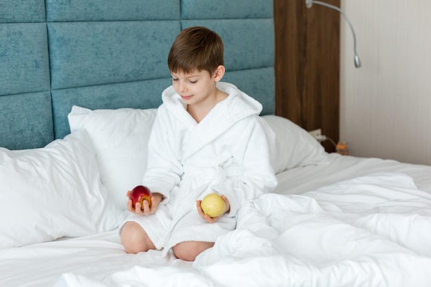 Un enfant en blouse blanche sur le lit sourit et tient des pommes Health Morning