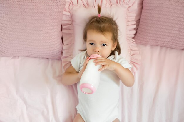 L'enfant boit un mélange de lait d'une bouteille se trouvant sur un lit avec des oreillers roses