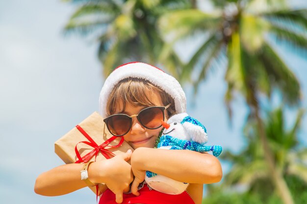 Enfant avec Bonnet de Noel et lunettes de soleil tenant des cadeaux à la plage
