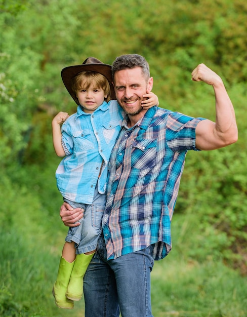 Enfant en bottes de caoutchouc papa heureux dans la forêt journée de la famille humaine et nature bonne journée de la terre Ferme écologique petit garçon aider père musclé dans l'agriculture père et fils en chapeau de cowboy sur le ranch plus forts ensemble