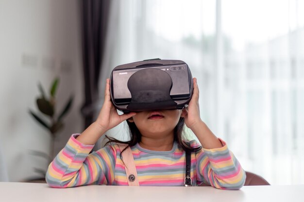 Enfant avec casque de réalité virtuelle assis derrière une table à l'intérieur à la maison