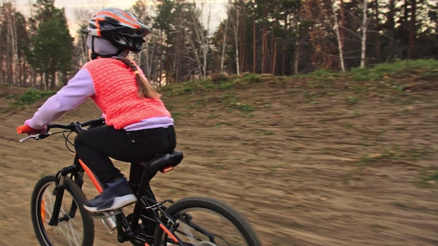 Un enfant caucasien chevauche une piste cyclable dans un parc de terre Fille faisant du vélo orange noir dans une piste de course Enfant va faire du vélo Sports Biker motion ride avec sac à dos et casque VTT hard tail