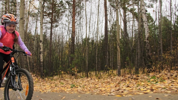 Un enfant caucasien chevauche une route cyclable dans le parc d'automne Petite fille faisant du vélo orange noir dans la forêt Enfant va faire du sport à vélo Biker motion ride avec sac à dos et casque VTT hard tail