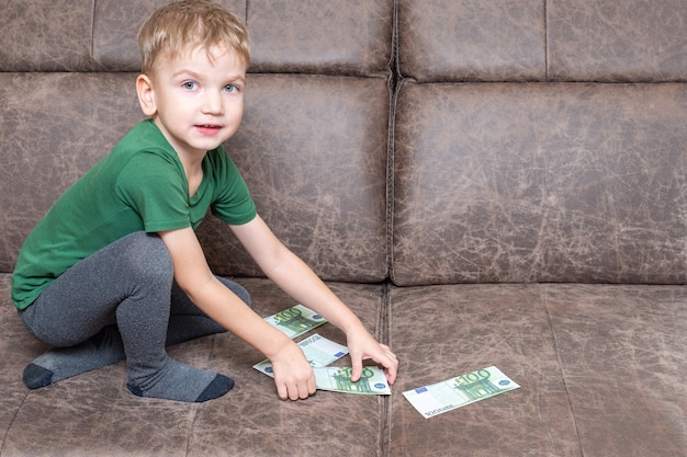 Un enfant caucasien concentré dans des vêtements décontractés tient cent euros dans ses mains et compte de l'argent sur le canapé de la pièce, vue latérale, espace de copie. L'éducation financière et économique des enfants