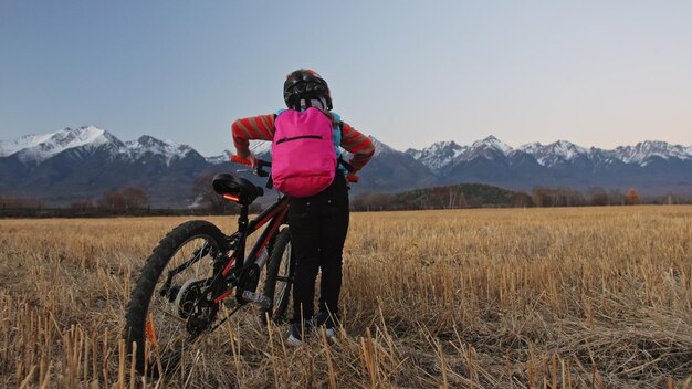 Un enfant caucasien marche avec un vélo dans un champ de blé Une fille qui marche un cycle orange noir sur fond de belles montagnes enneigées Biker motion ride avec sac à dos et casque VTT hard tail